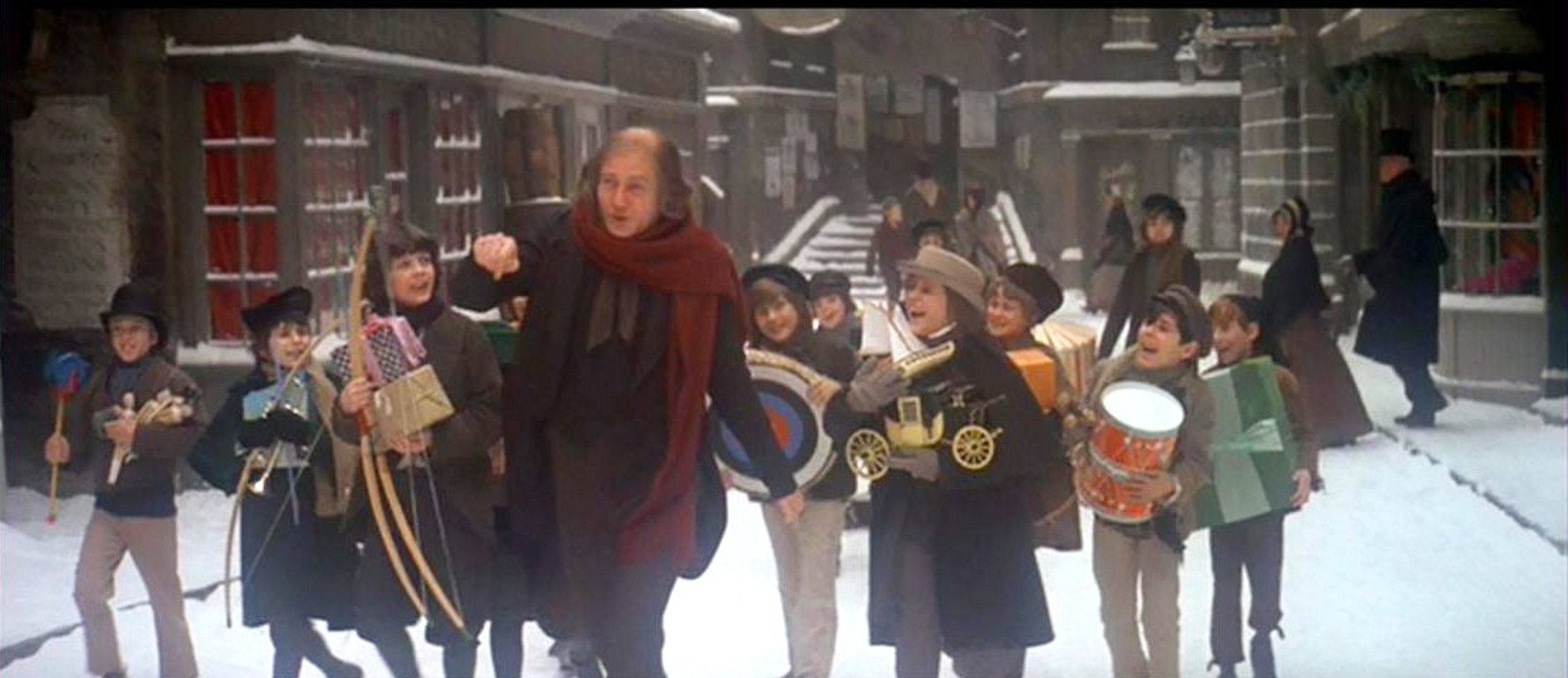Still from the 1970 film. Scrooge happily leads a group of children down a snowy street.