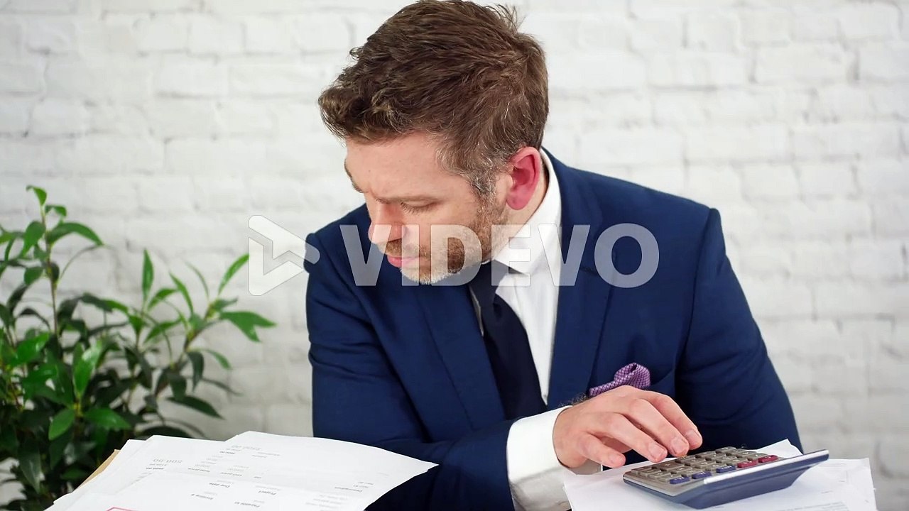A stressed businessman using a calculator calculating debts and taxes with piles of financial accounts paperwork in an office
