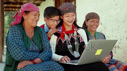 A cheerful Asian Indian group of rural family members including mixed aged women wearing traditional costumes and a kid smiling and watching funny online movie or video on a laptop in a remote village