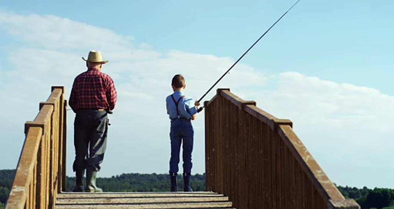 Rear view of a grandfather fishing with his little grandson while standing on the wooden bridge and holding rods in the water