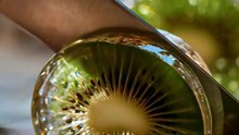 A hand holding a knife cutting a kiwi fruit looks like clear glass on a wooden board.