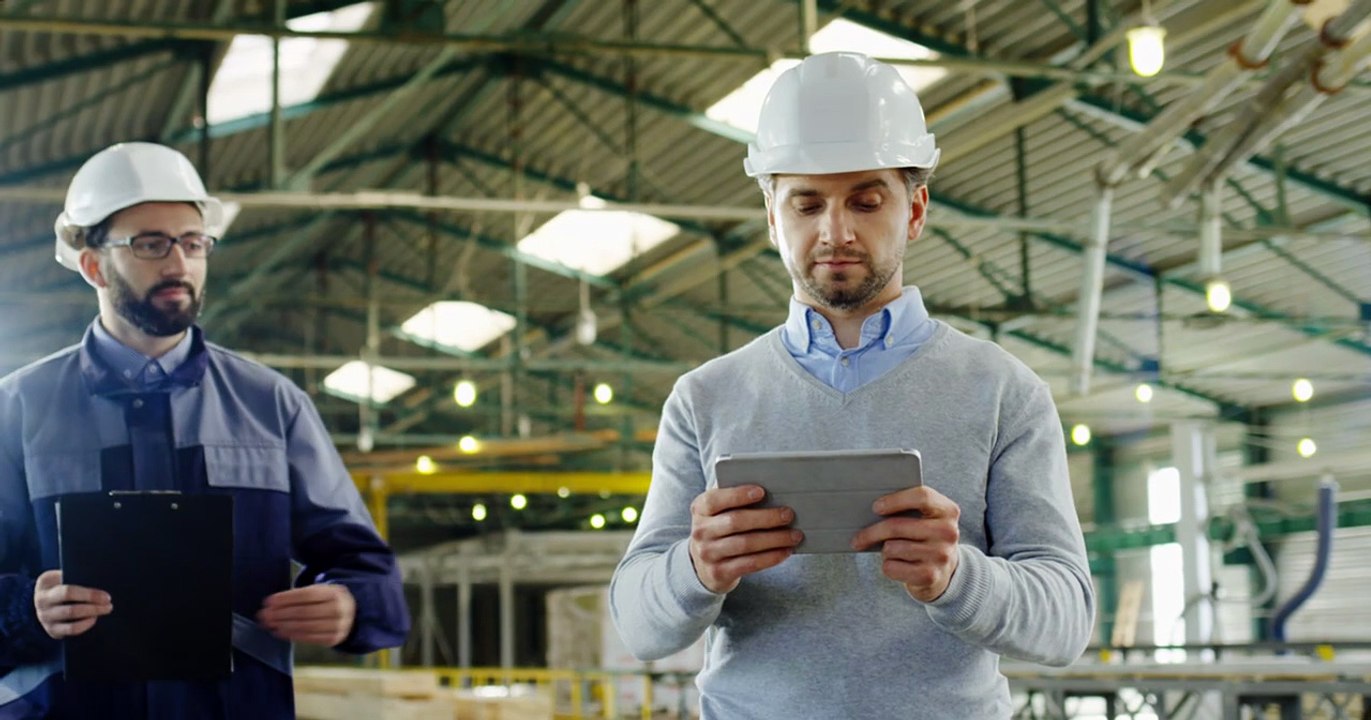 caucasian male worker wearing a helmet and using a tablet in a factory, then his coworker comes up and talking to him