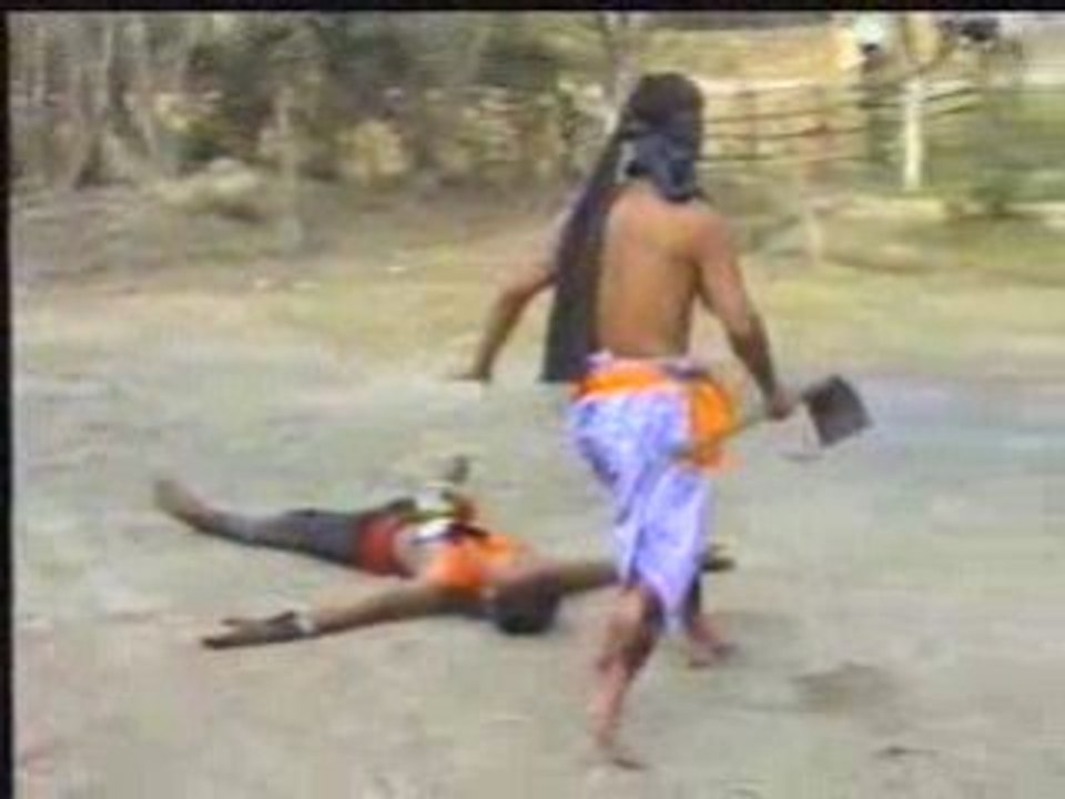 blindfolded man uses hoe to cut a papaya placed on another man's stomach
